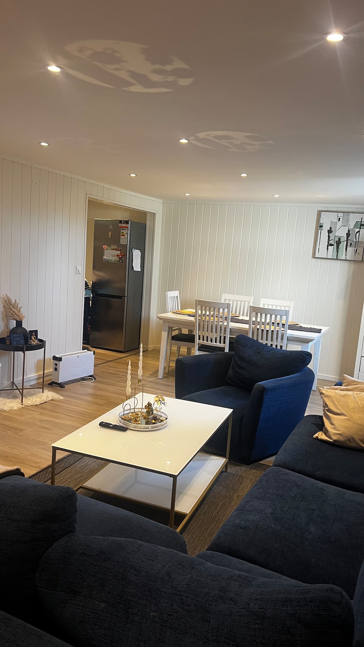 A living area features a dark blue sofa and a light-colored coffee table, providing a cozy atmosphere. In the background, a dining table with white chairs is visible, along with a sleek stainless steel refrigerator. Soft lighting enhances the neutral-colored walls.