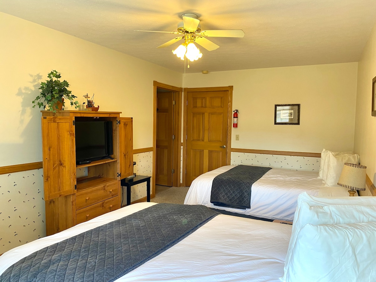 A bright bedroom features two spacious beds dressed with white linens and dark grey quilts. A wooden TV cabinet is positioned against the wall, complemented by a small table. Natural light enters through a nearby window, enhancing the overall ambiance of the room.