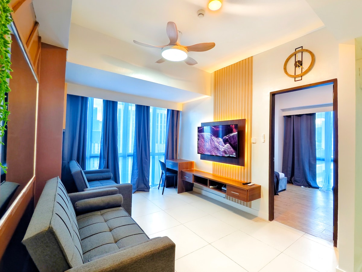The living area features a modern design with two gray sofas and a wooden entertainment unit housing a large flat-screen TV. Natural light filters through the large windows, which are dressed in dark curtains, complementing the neutral tones of the space.