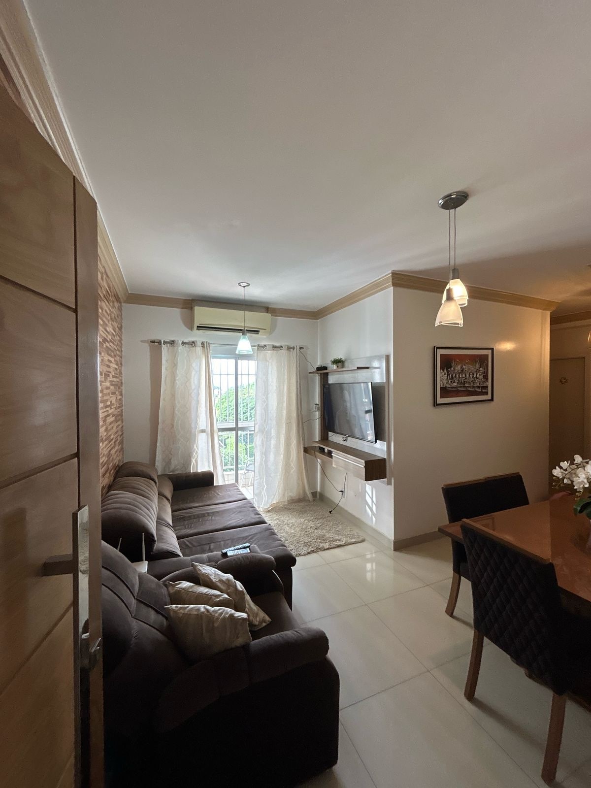 A light-filled living area showcases a comfortable sectional sofa and a modern entertainment unit. A dining table is set against a neutral wall, while large windows draped with sheer curtains allow natural light to illuminate the space. Decorative elements, like a potted plant, provide a welcoming touch.
