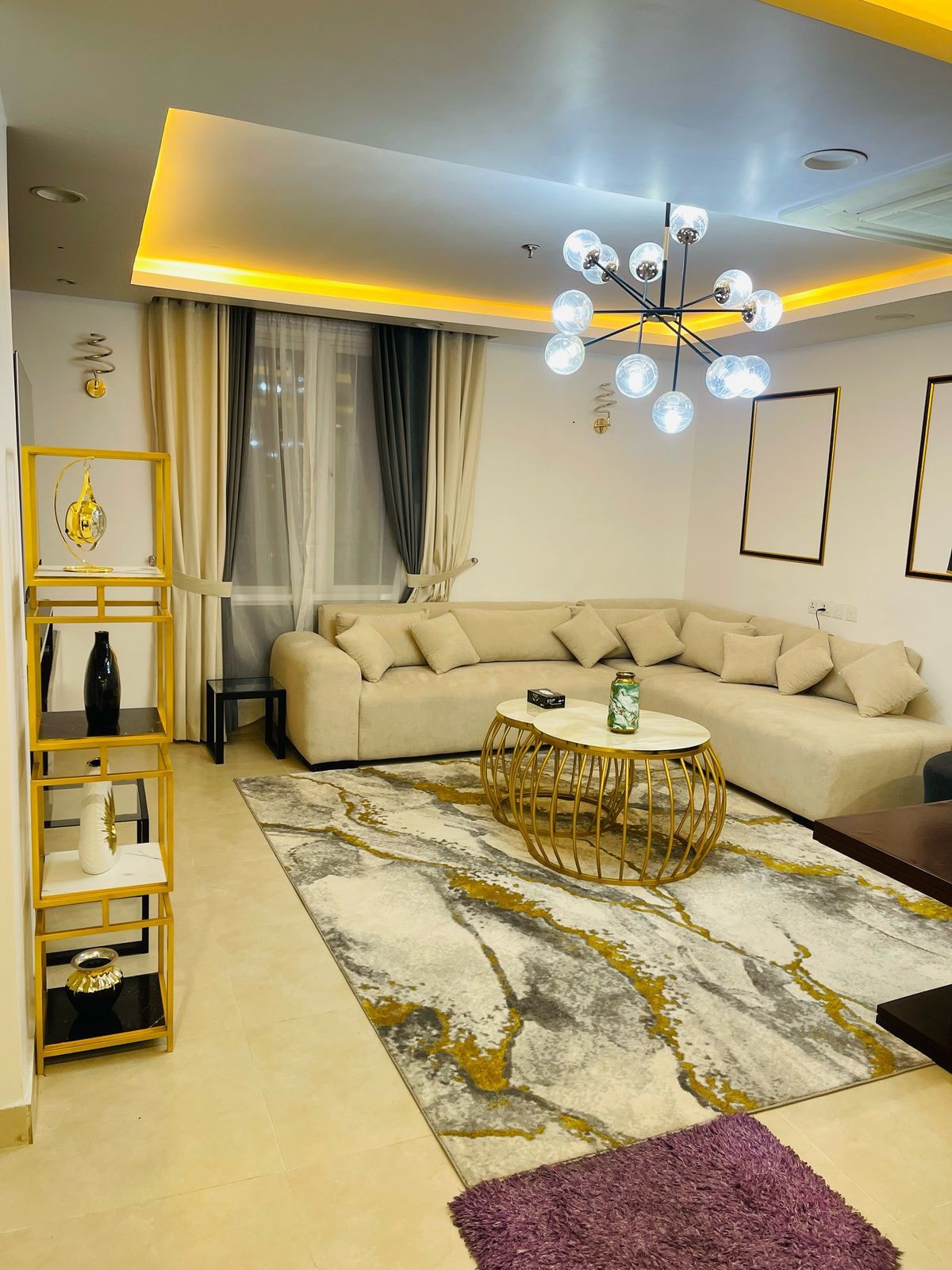 A spacious living area features a large, light-colored sectional sofa arranged around a round coffee table. A patterned area rug with gold accents adds warmth to the space, while soft lighting illuminates the room from a modern chandelier hanging above.