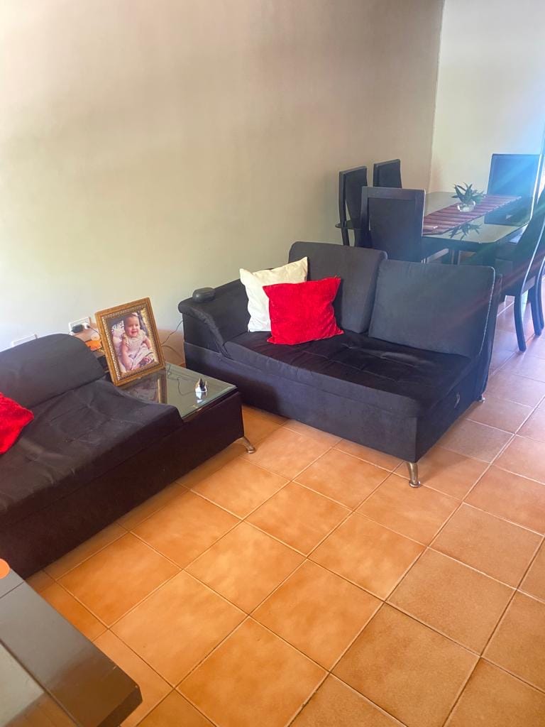 A comfortable living area features two black sofas adorned with red and white cushions. A glass coffee table sits between them, and a framed photograph is positioned on the floor next to the table. In the background, a dining table with several chairs is visible.