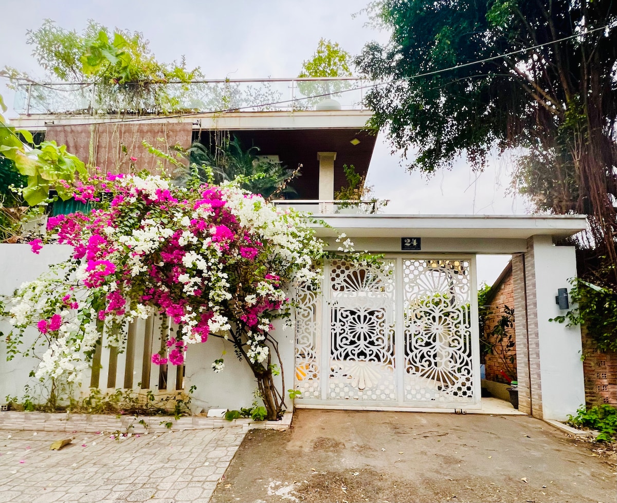 The entrance is marked by a decorative gate adorned with intricate patterns. Surrounding foliage includes blooming bougainvillea in vibrant pink and white, contributing to a welcoming atmosphere. Above, a terrace can be seen, adding an inviting layer to the exterior.