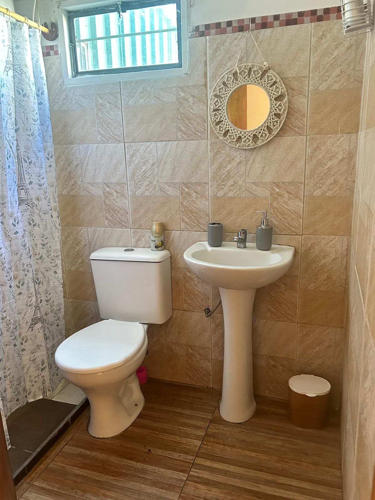 An inviting bathroom is featured, with a white toilet and a pedestal sink. A large mirror enhances the space, while soft hues of the tiled walls create a calming ambiance. Natural light filters in through a small window, complemented by a patterned shower curtain.