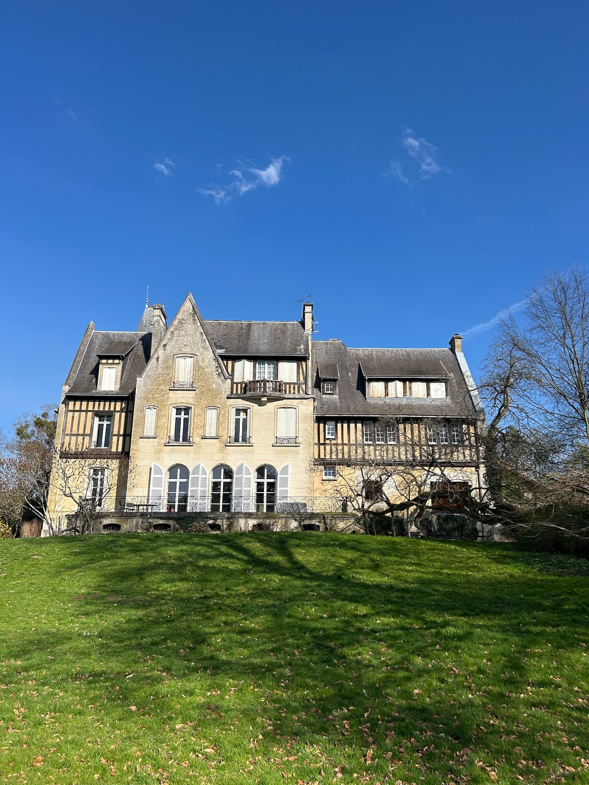 The exterior of the house is depicted against a clear blue sky, showcasing a historic architectural style with pointed roofs and multiple windows. A grassy expanse leads to the building, surrounded by trees that provide natural shade.