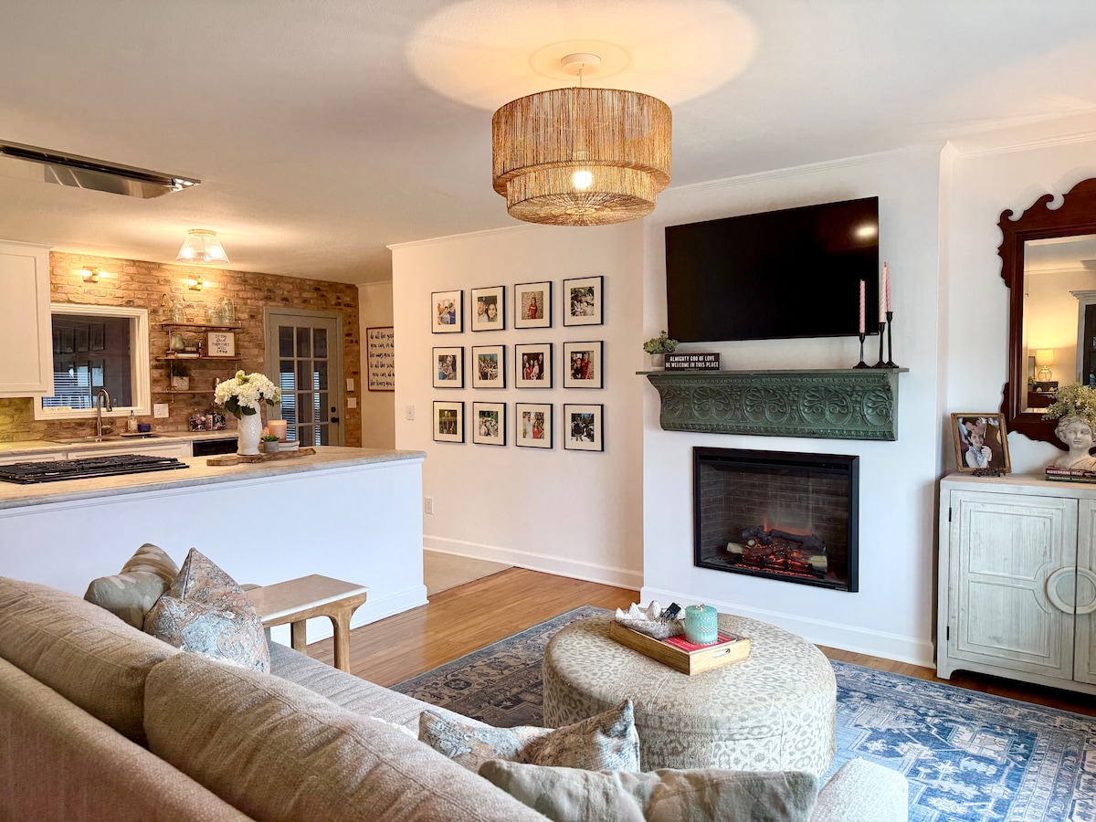 The living area features a comfortable sofa with decorative pillows, a circular coffee table, and a stone fireplace adorned with a mantel. A flat-screen television is mounted above the fireplace, and a stylish light fixture hangs from the ceiling, providing a welcoming ambiance.