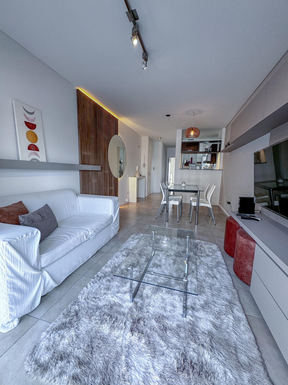 An open living space features a white sofa adorned with colorful cushions. A glass coffee table sits on a plush grey rug, while a small dining area is highlighted by a round table and transparent chairs. Subtle lighting accentuates the modern decor.