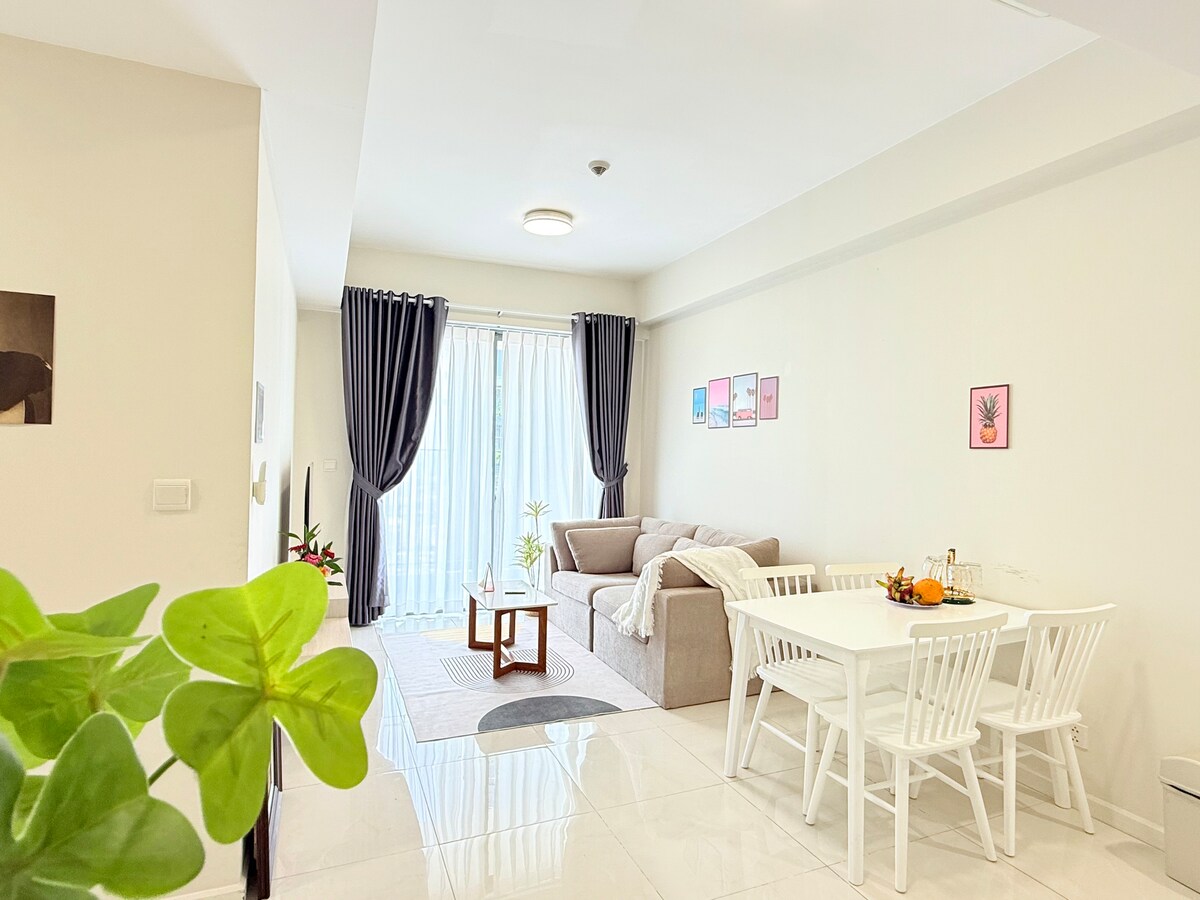 A bright living area is featured, showcasing a comfortable sofa and a coffee table. A dining table with white chairs is positioned beside large glass doors, leading to a balcony. Decorative wall art adds a pop of color to the neutral-toned walls.