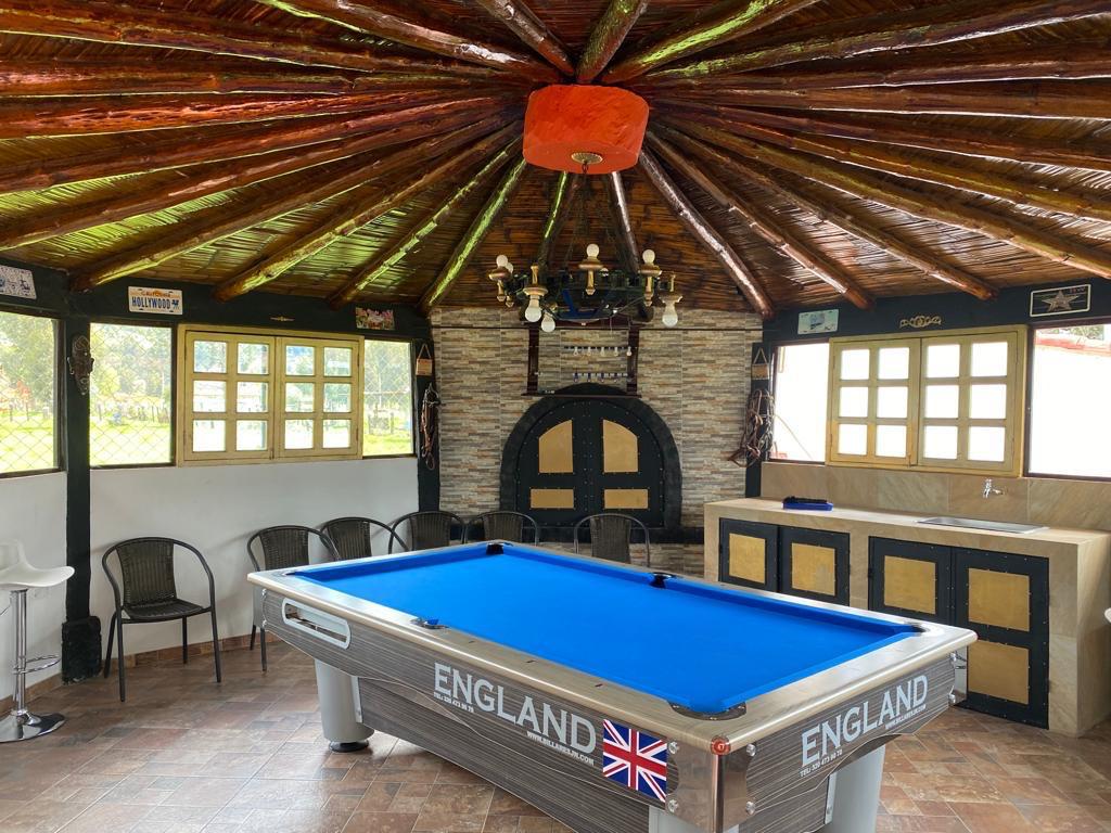 A recreational area features a billiards table with a blue felt surface, surrounded by several chairs. Natural light enters through multiple windows, while a stone fireplace adds a rustic touch. The wooden ceiling showcases a unique design, creating an inviting space for relaxation.