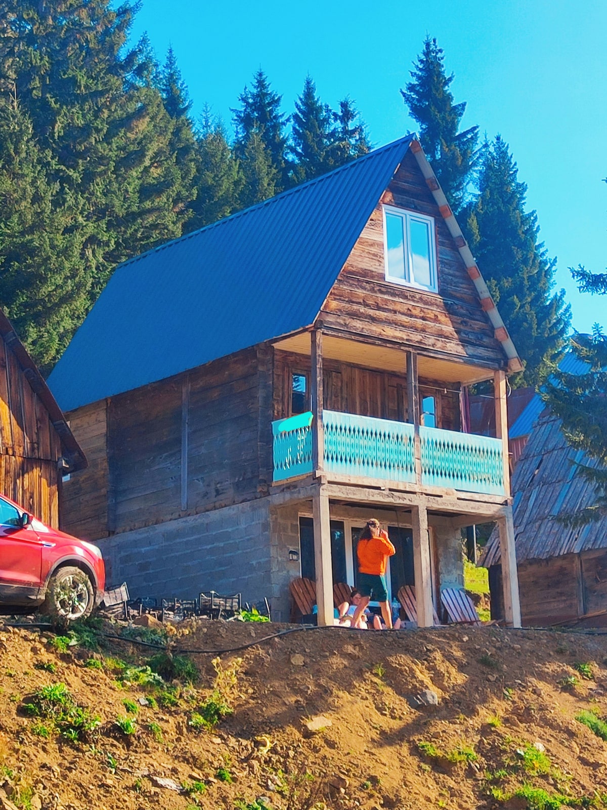 A charming cabin constructed with rustic wooden materials is displayed from a hillside perspective. The cabin features a balcony with a light blue railing and large windows, surrounded by tall conifer trees. A red vehicle is parked nearby, indicating accessible amenities.
