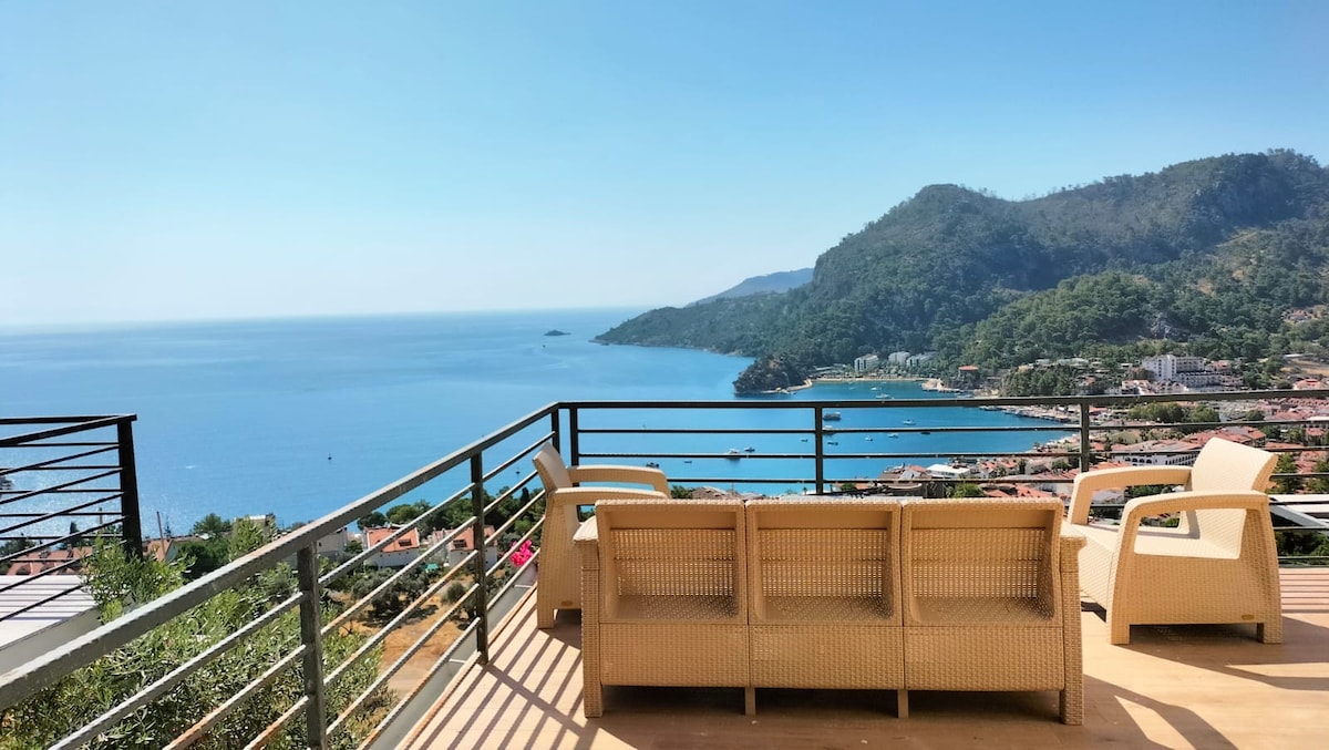 A spacious outdoor terrace is shown with comfortable woven seating arranged to face a scenic view of the sea. The mountains and coastline are visible in the background, under a clear blue sky, creating an inviting space for relaxation.