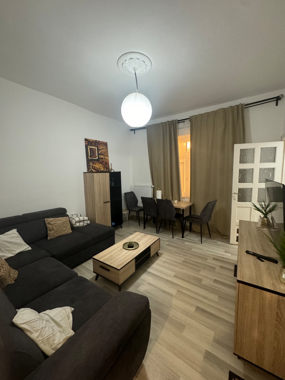 A comfortable living area features a dark sectional sofa and a wooden coffee table at its center. A dining space with six chairs is visible in the background, alongside a window partially covered by light curtains. A pendant light hangs from the ceiling, providing soft illumination.