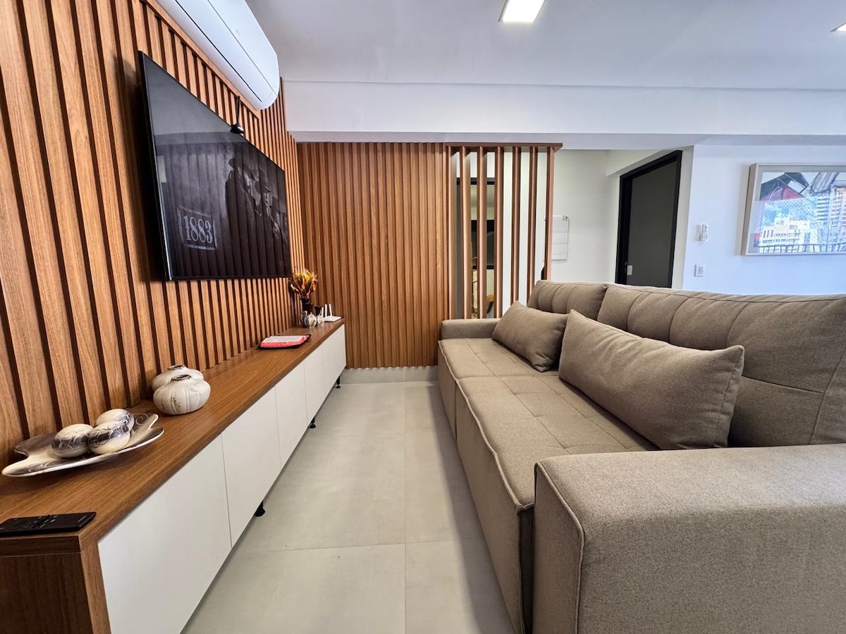 A modern living area features a spacious, light-colored sofa positioned in front of a wall-mounted television. Wooden paneling creates a warm backdrop, while a sleek console holds decorative items. Natural light brightens the room through large open spaces, enhancing the contemporary design.