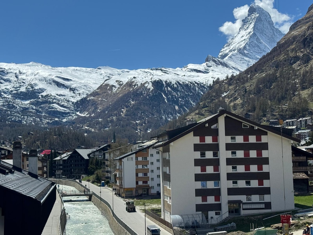 Snow-capped mountains are visible against a clear blue sky, with the distinct peak of the Matterhorn in the background. A river runs along the road in the foreground, flanked by modern apartment buildings, creating a scenic view of the surrounding landscape.