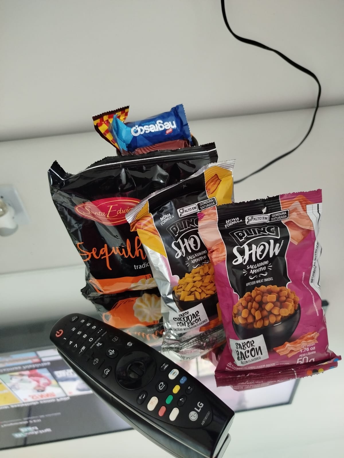 An assortment of snack bags is displayed on a reflective surface. The snacks featured include a chocolate bar and various popcorn options. A remote control is positioned in front, with visible buttons for easy accessibility, while a screen showing entertainment content is reflected in the background.