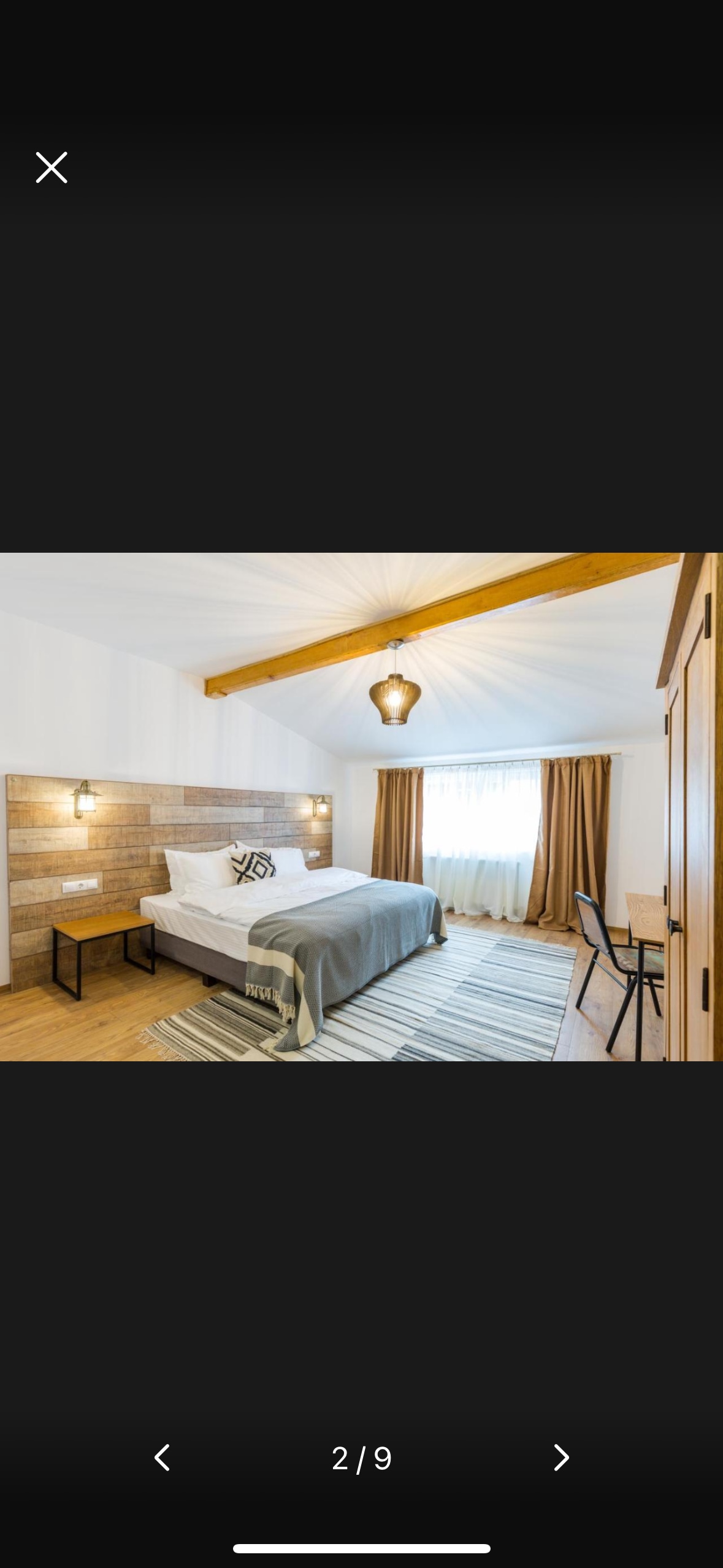 A spacious bedroom is presented with a large bed featuring a layered blanket. Wooden beams enhance the ceiling, while natural light fills the room through large windows covered by light brown curtains. A desk and two chairs are positioned near the corner against a neutral wall.