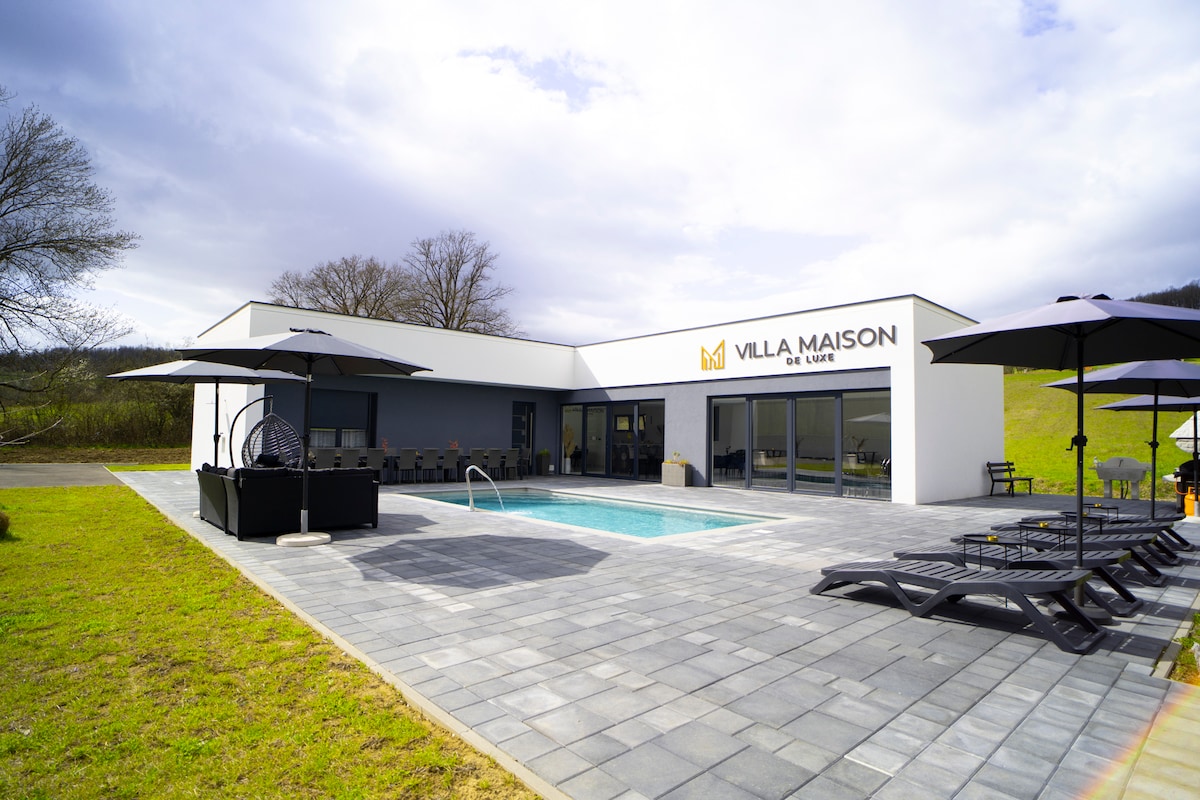 A modern villa is shown with a clean architectural design. An inviting swimming pool is set in the foreground, surrounded by lounge chairs. Dark umbrellas provide shade on the terrace, while the exterior features large windows reflecting the surrounding landscape.