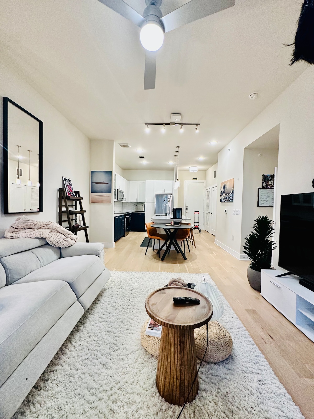 An open living area is featured, showcasing a light-colored sofa positioned next to a round wooden coffee table on a soft rug. A dining table with orange chairs is visible in the background, along with a modern kitchen fitted with stainless steel appliances.