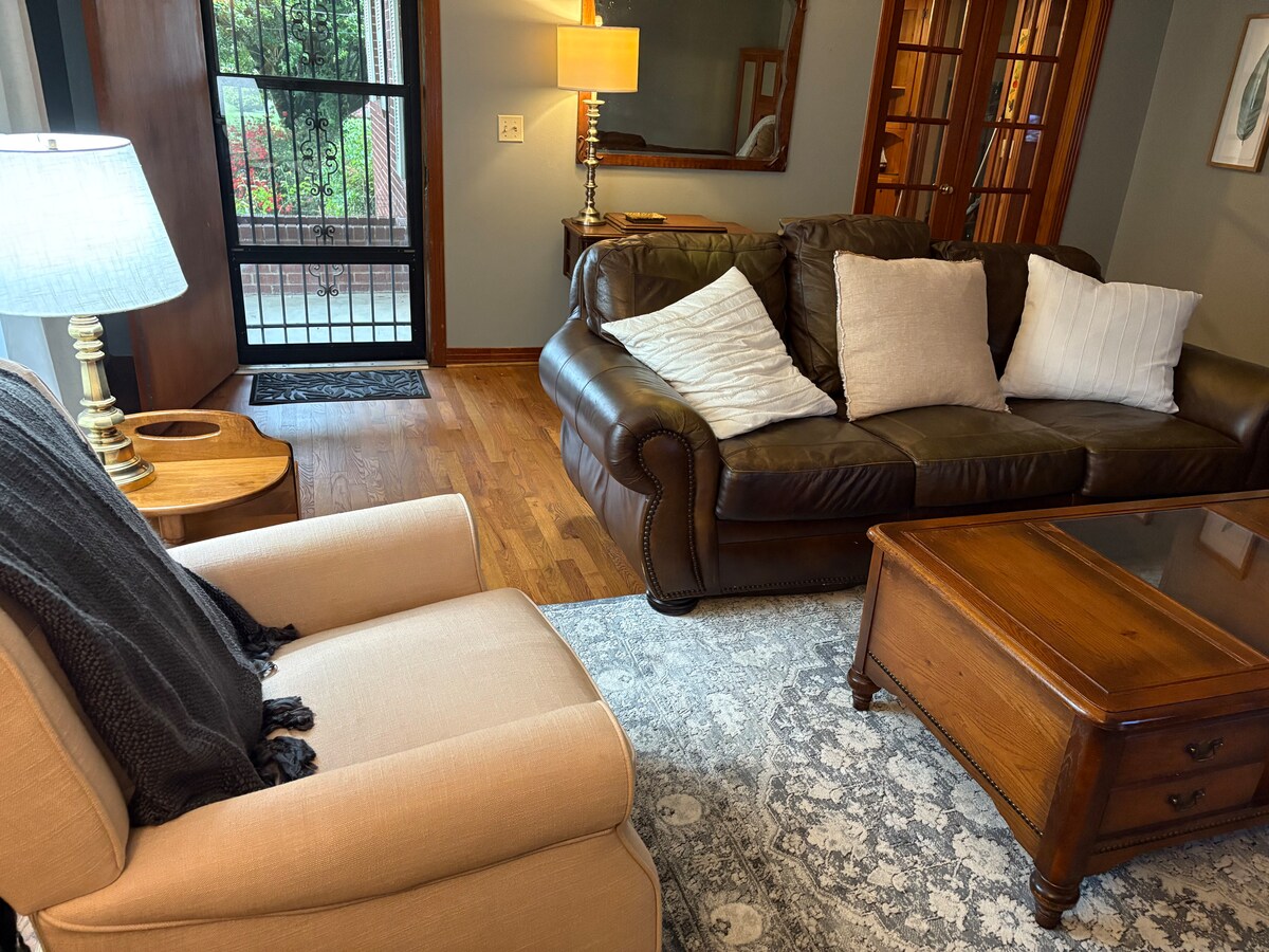 A cozy living area features a dark leather sofa adorned with white pillows, positioned alongside a light armchair. A wooden coffee table sits in front, resting on a decorative rug. Soft light emanates from a nearby lamp, complementing the ambiance.