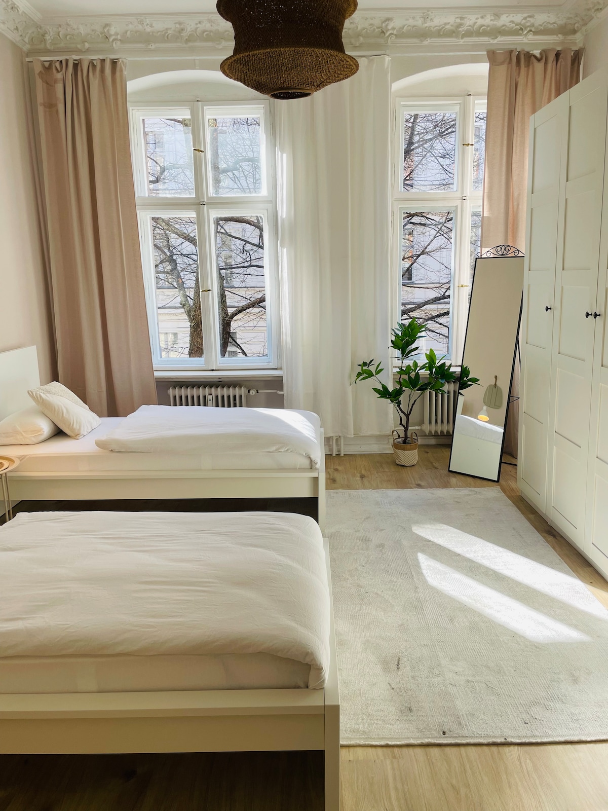 A bright bedroom is presented, featuring two spacious beds with white bedding. Natural light streams through tall windows adorned with light curtains. A plant is positioned near a full-length mirror, complementing the neutral tones of the room. A soft rug adds warmth to the wooden floor.