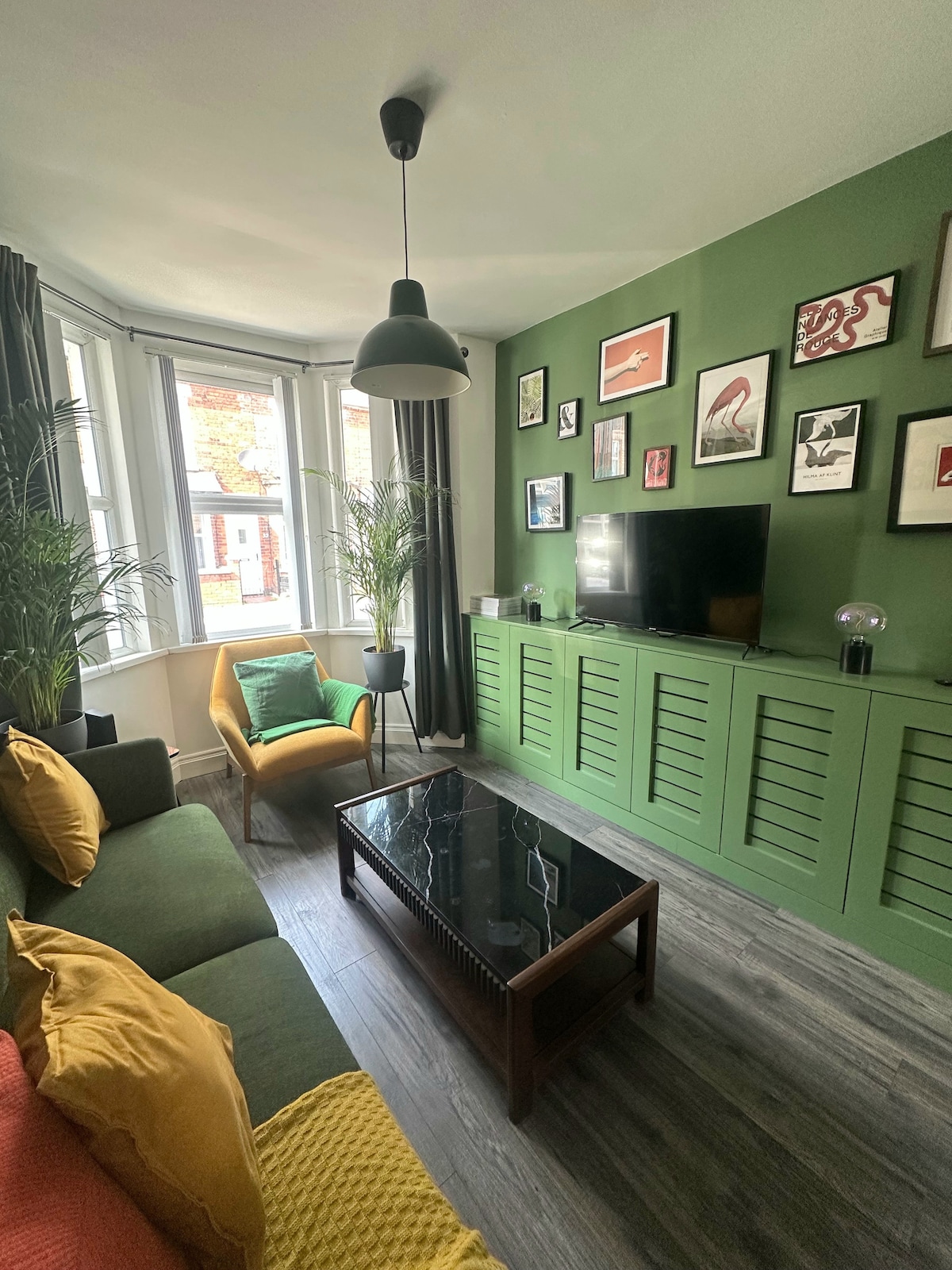 A cozy living area features green walls and large windows allowing natural light to fill the space. A green sofa with yellow cushions sits alongside a bright accent chair. A sleek black coffee table is centered on the floor, complemented by decorative wall art and indoor plants.