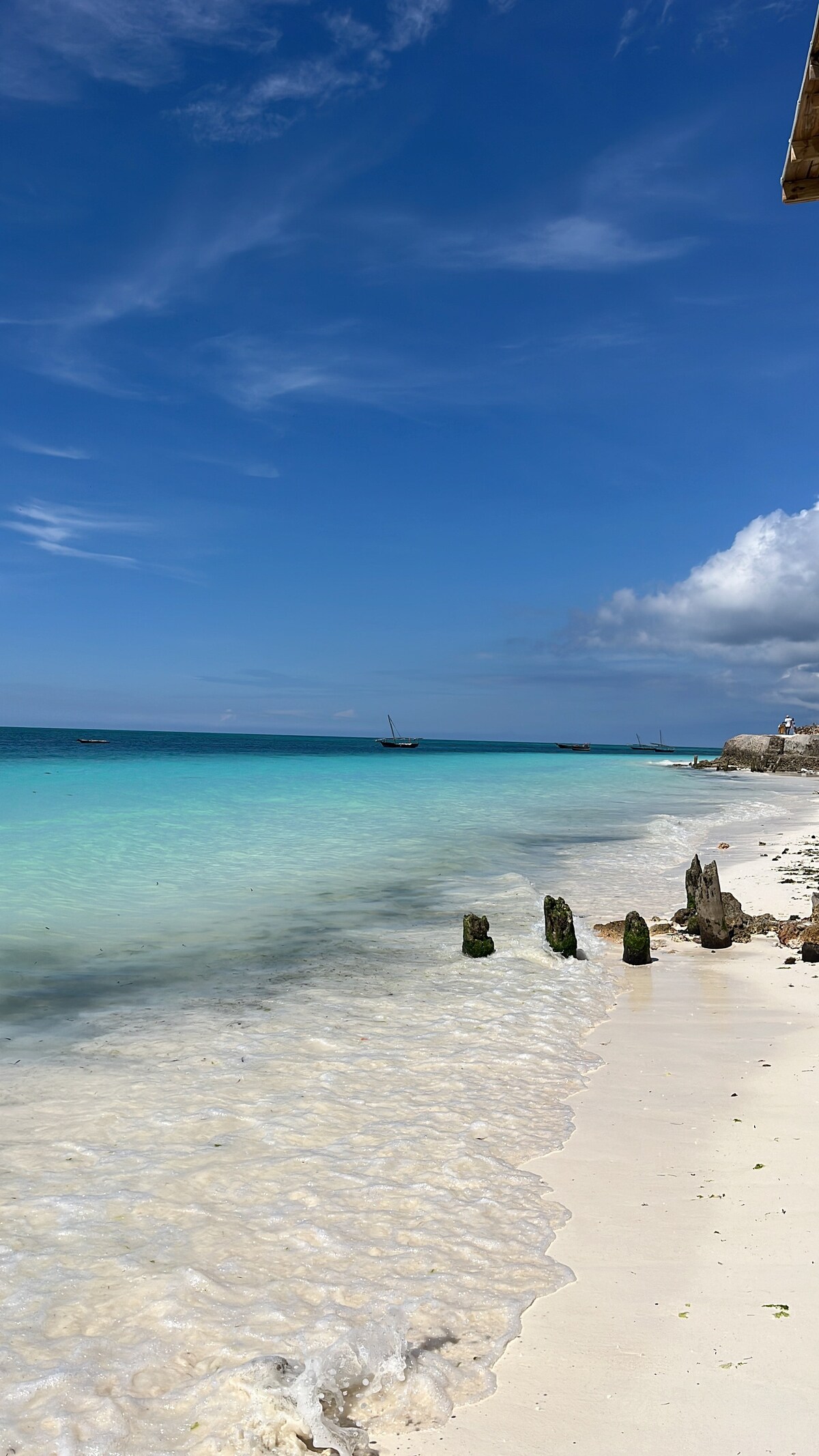 A serene beach scene showcases soft white sand meeting clear turquoise waters. Gentle waves lap at the shore, while a few fishing boats are visible in the distance. A partly cloudy sky adds dimension to the tranquil setting.