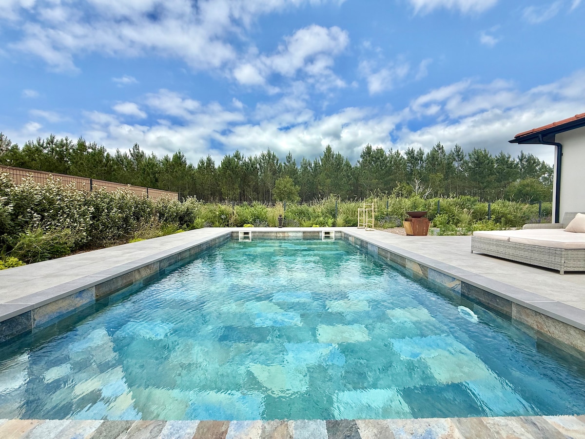 A serene pool area is surrounded by lush greenery, featuring clear water reflecting the sky. A comfortable seating arrangement is visible on the poolside, along with a landscaped area designed for relaxation and enjoyment.