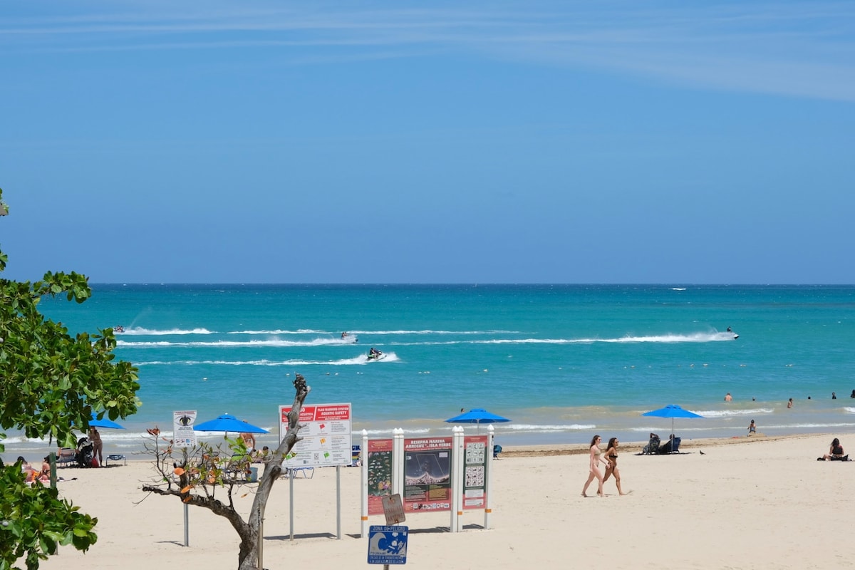 A sandy beach extends before the viewer, framed by vibrant blue umbrellas and a clear turquoise ocean. Jet skis create gentle waves in the water, while a few people enjoy the sun along the shoreline. Informational signs and a tree are visible, adding to the beach's inviting atmosphere.