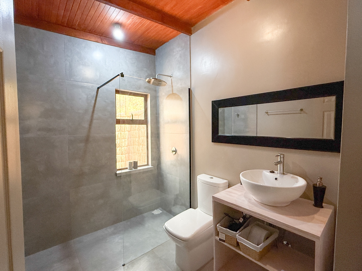A modern bathroom is highlighted, featuring a spacious shower with a glass enclosure. A round vessel sink sits atop a light wood vanity, accompanied by neatly organized storage options. Soft lighting complements the warm tones of the wood ceiling and the textural wall finishes.