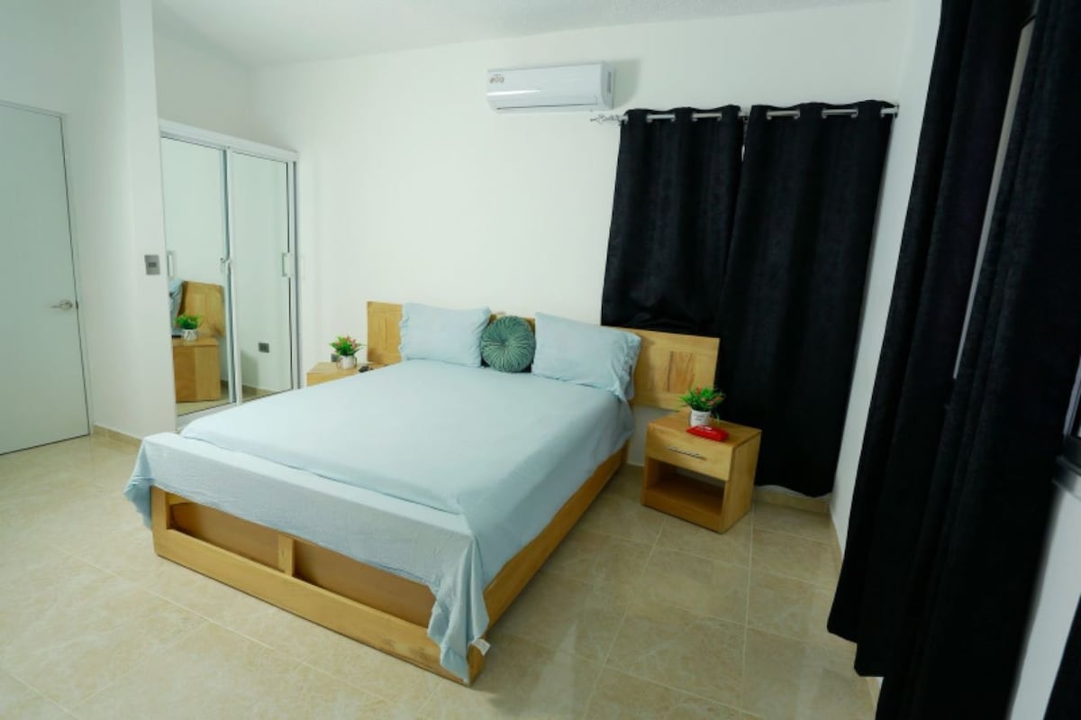 A bright sleeping area features a spacious bed with a light blue coverlet, flanked by two nightstands. A large mirror cabinet reflects the room's neutral tones, while dark curtains frame the window. Indoor plants add a touch of greenery to the serene environment.