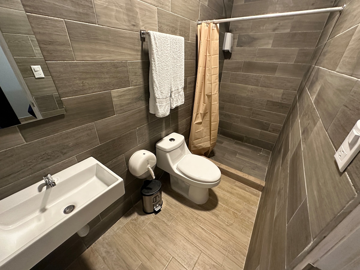 A modern bathroom features tiled walls in neutral tones. A spacious shower area is present, with a beige curtain for privacy. A compact sink and toilet are visible, along with a towel rack holding two white towels. A small waste bin completes the setup.