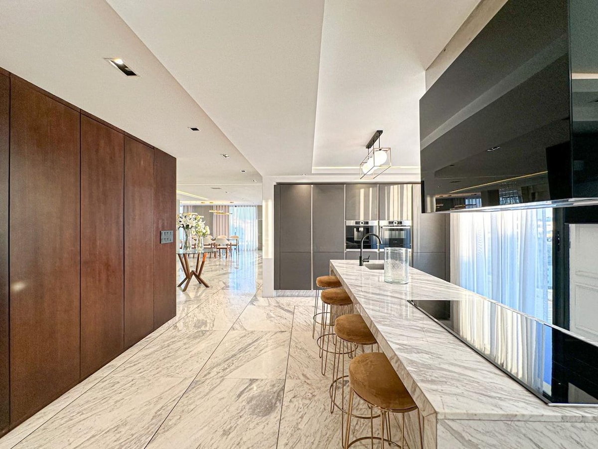 A spacious kitchen area is highlighted by a marble island with bar seating. Dark cabinetry and modern appliances create a sleek design, while natural light floods the space through large windows. The open layout connects seamlessly to the dining area in the background.