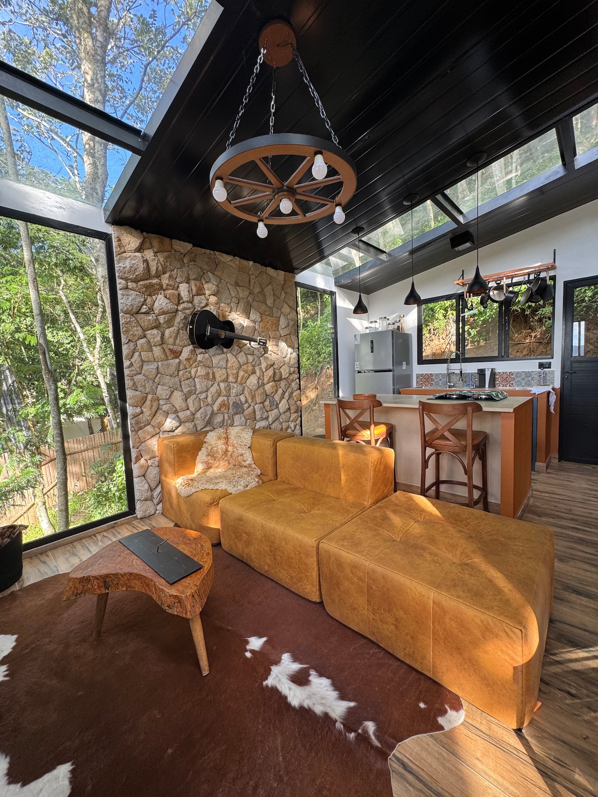 A bright living area is showcased with large windows offering views of greenery. A spacious, modular brown sofa is complemented by a rustic wooden coffee table. The room features a stylish stone accent wall and a modern kitchen space in the background, illuminated by pendant lighting.