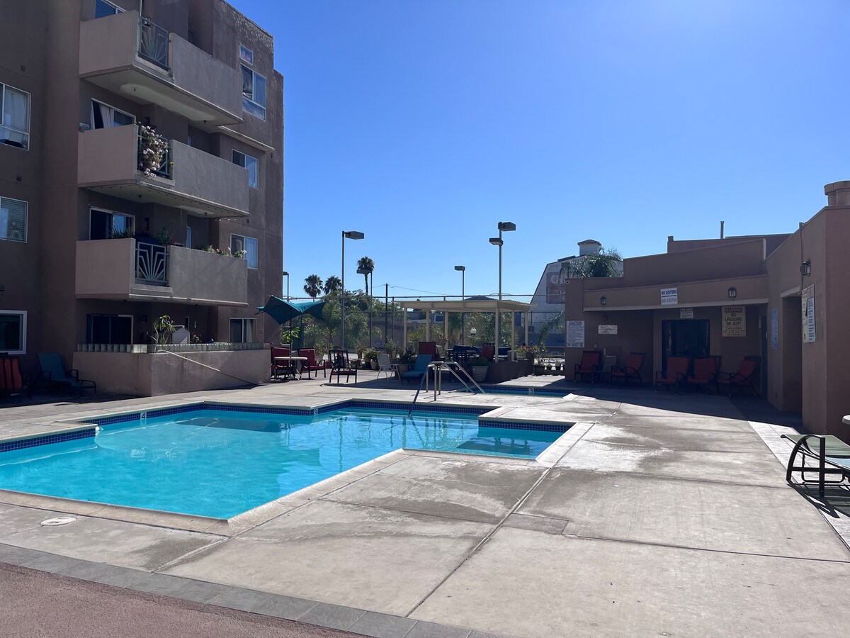 A community pool is highlighted, surrounded by a spacious deck with lounge chairs for relaxation. Nearby, a shaded seating area is visible. The bright sky and sunlight create a warm, inviting environment, enhancing outdoor leisure opportunities.