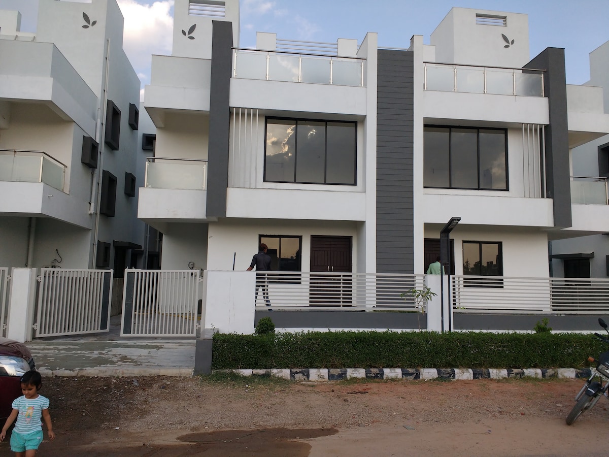 A modern two-story building is displayed, featuring a combination of white and gray exterior elements. Balconies are present on the upper levels, with large windows allowing natural light. A neatly landscaped area with grass borders the property, and a secure gate is visible at the entrance.