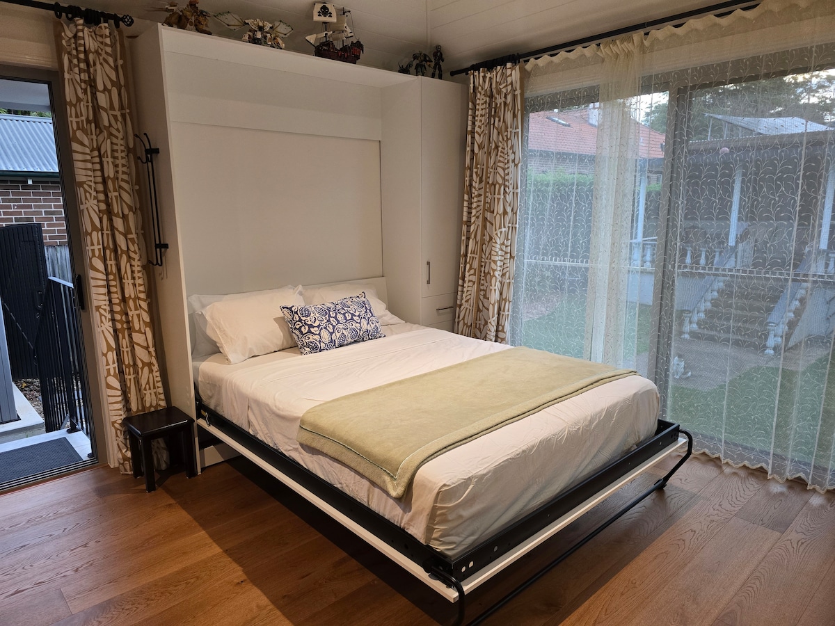 A cozy sleeping area features a queen-sized wall bed pulled down, adorned with a decorative blue pillow. Light beige curtains frame a large window that provides a view of the backyard, while wooden flooring adds warmth to the space.