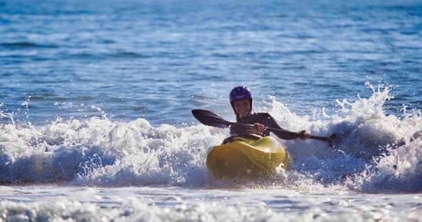 A person is maneuvering a kayak through gentle ocean waves. Sunlight glimmers on the water's surface, creating a lively scene. The surrounding sea reflects a clear blue sky, providing a vibrant backdrop for the water activity.