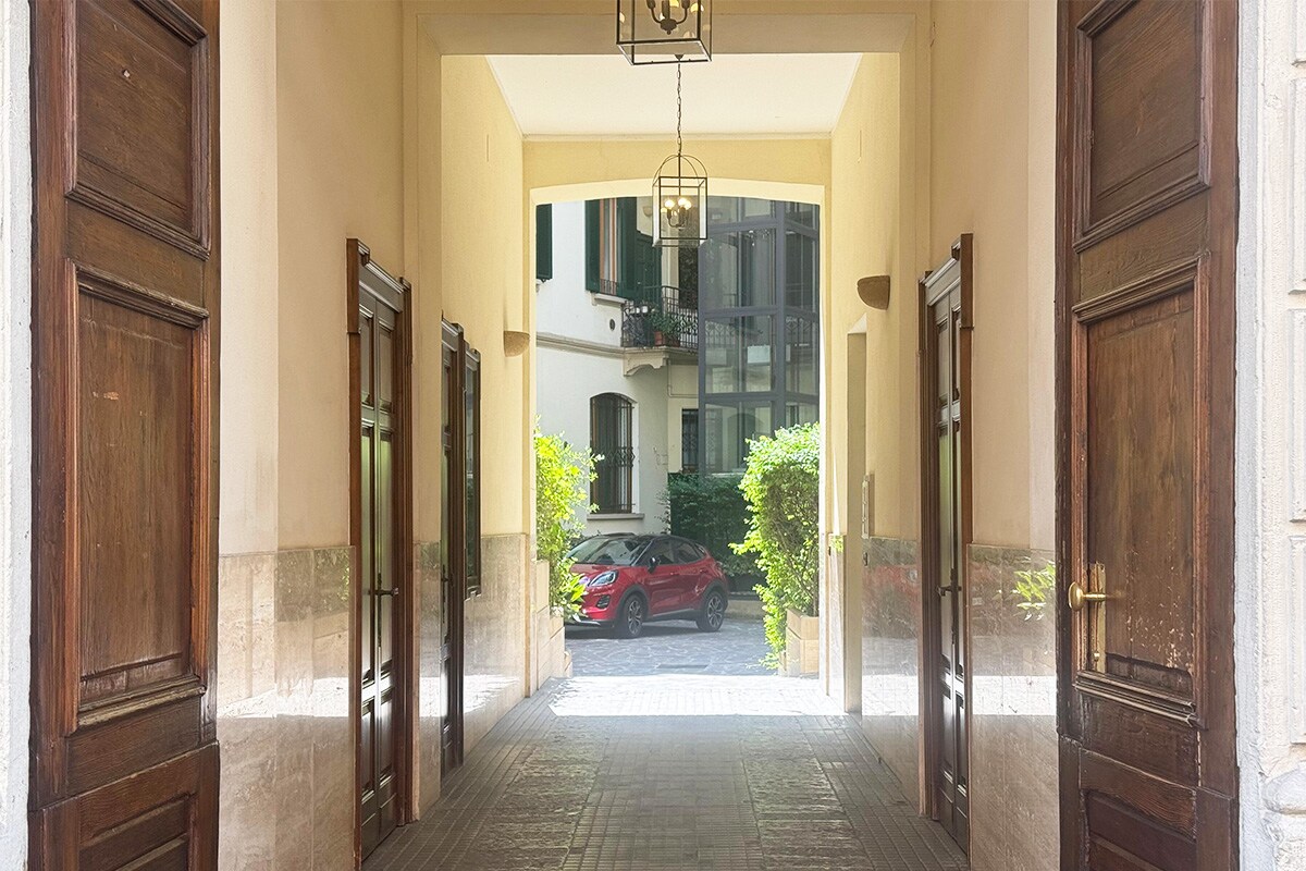 An elegant entrance is framed by wooden doors and features a passageway leading to a courtyard. Lush greenery is visible along the sides, while an inviting car is parked at the end, enhancing the sense of accessibility and tranquility.