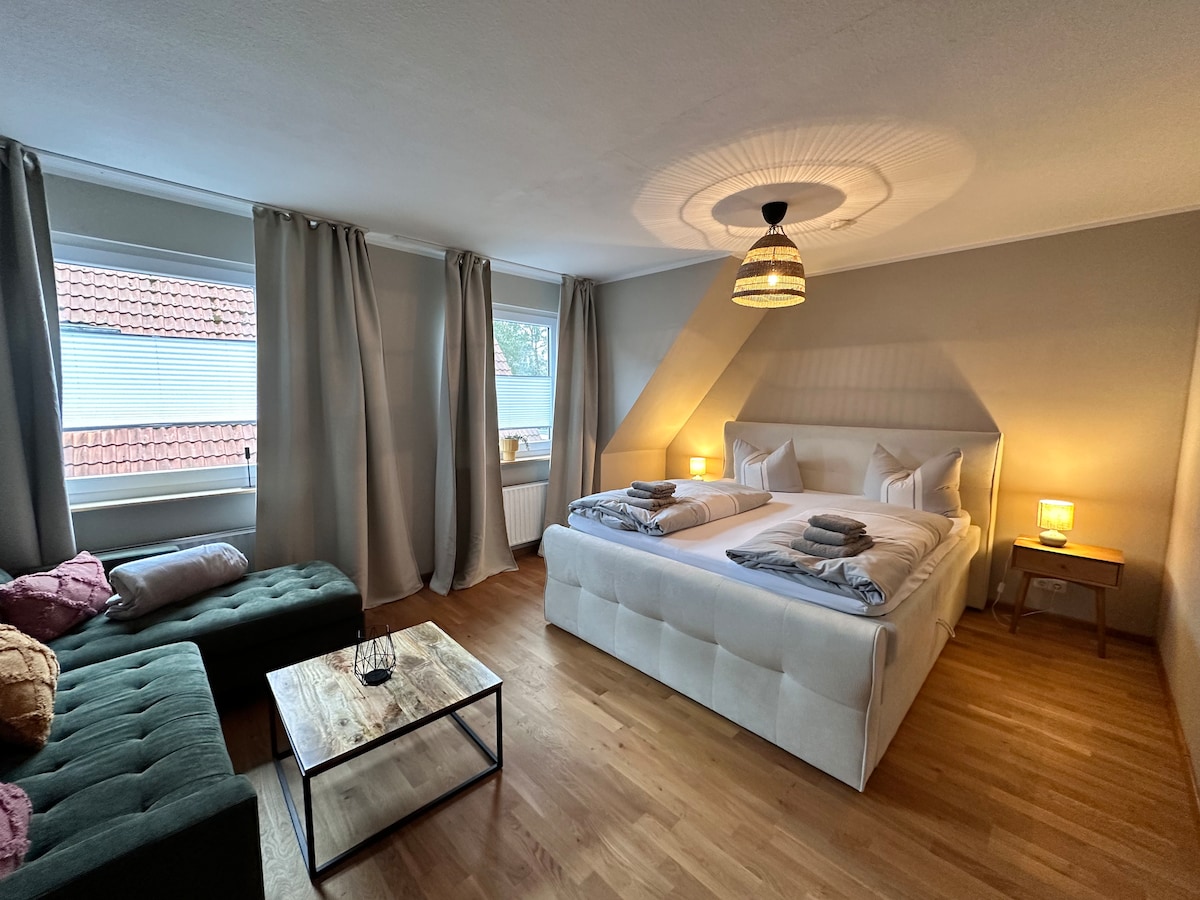 A spacious bedroom features a large bed dressed with neutral linens and two white pillows. A dark green sofa with decorative cushions sits beside a wooden coffee table. Soft light is provided by lamps on either side of the bed, and natural light filters through two windows with curtains.