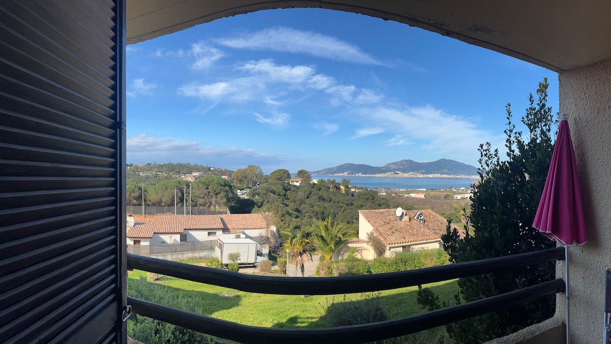 A scenic view is captured from a balcony, showcasing rolling hills and the coastline in the distance. Clear blue skies and a hint of greenery frame the landscape. A pink umbrella is visible, adding a pop of color to the neutral tones of the surroundings.
