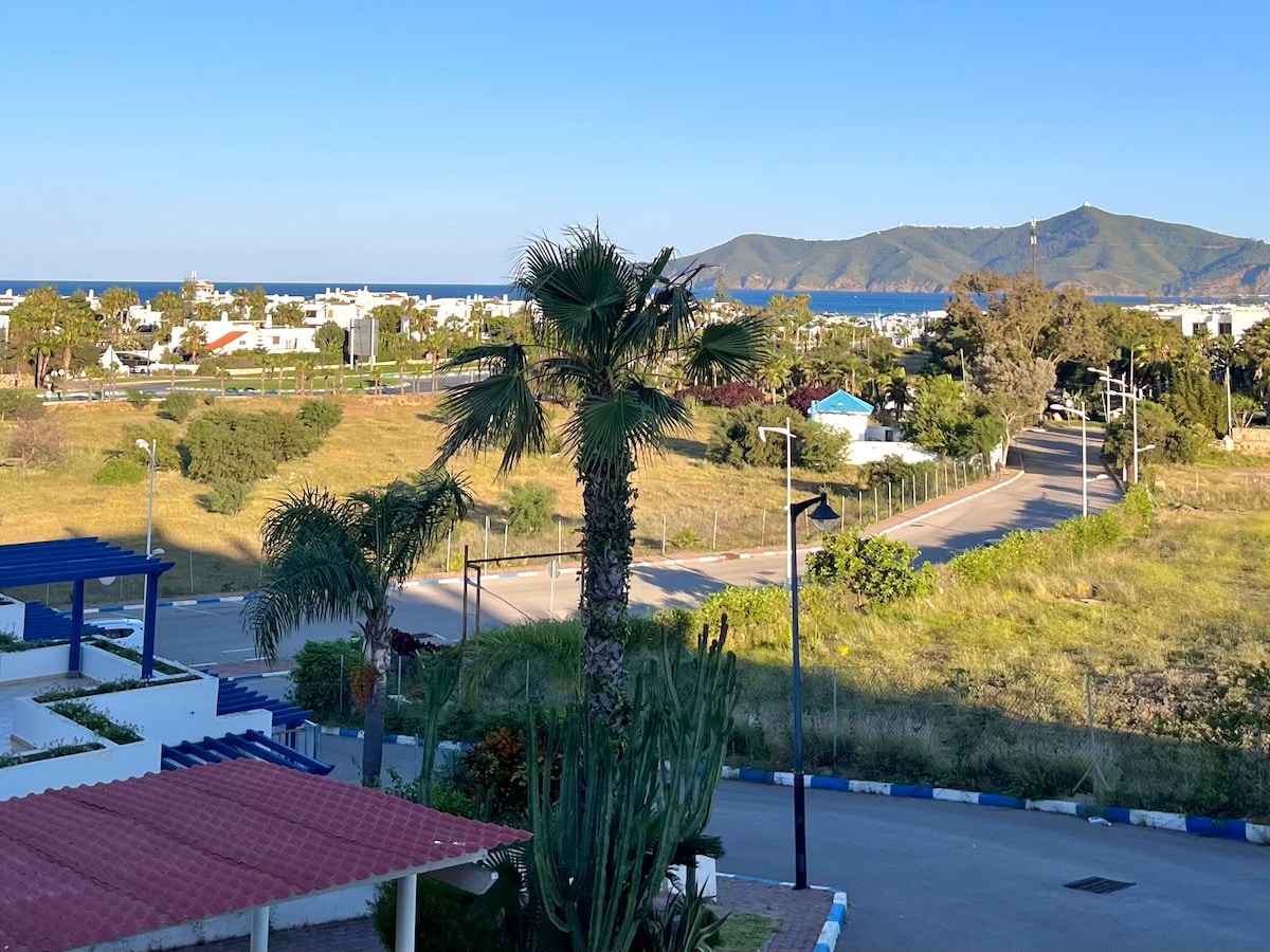 A scenic view from a terrace captures the lush greenery and palm trees, with the coastline visible in the distance. Rolling hills extend towards the horizon, framed by a clear blue sky, while a peaceful road meanders among the well-maintained surroundings.