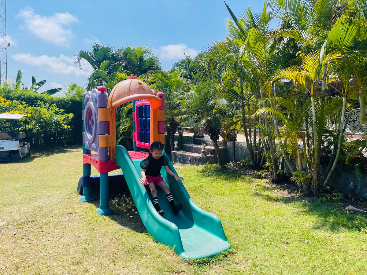 A colorful playground structure is situated on a sunny lawn, featuring a slide and climbing areas. Surrounding palm trees provide shade, and the clear blue sky enhances the outdoor setting.