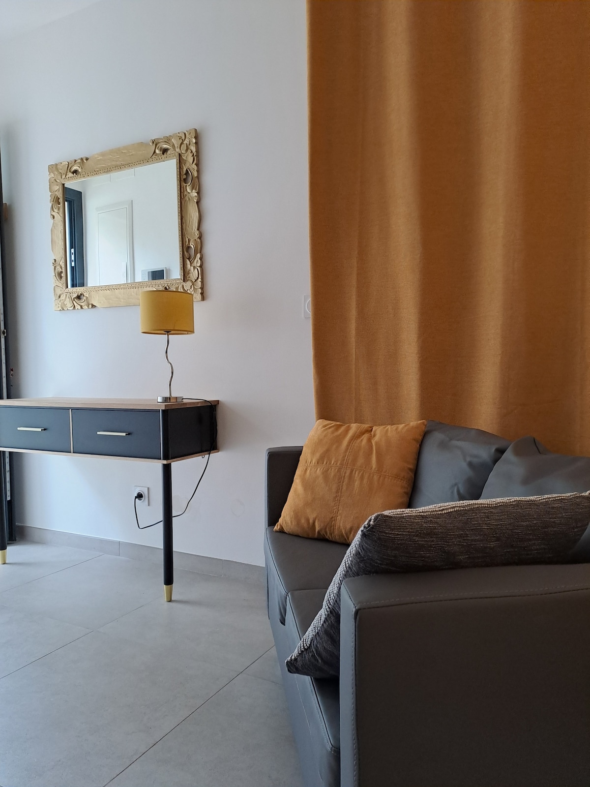 A contemporary living area features a sleek grey couch accented by decorative cushions. A stylish lamp rests on a dark side table, complementing a gold-framed mirror mounted on the wall. Soft, warm drapes enhance the inviting atmosphere, while natural light filters through nearby windows.