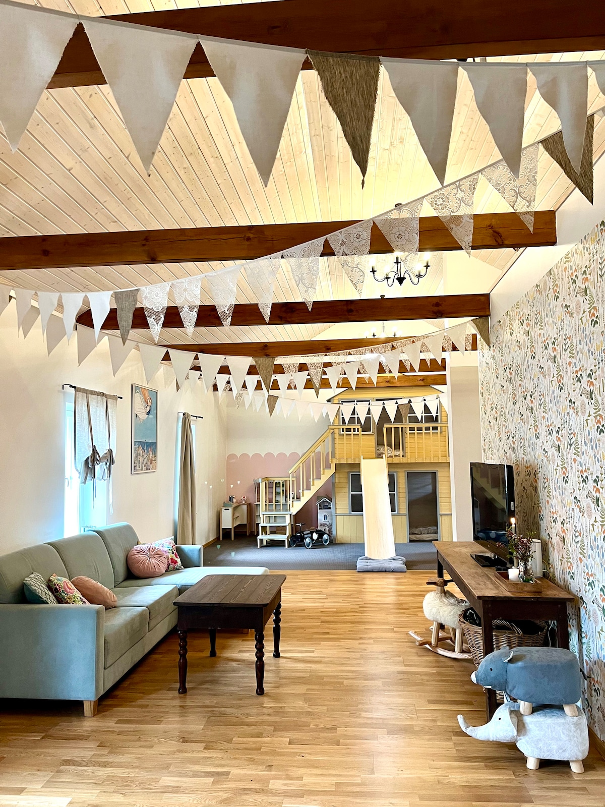 A spacious living area features a light wooden ceiling with exposed beams, adorned by decorative bunting. A soft couch and a wooden coffee table are positioned on warm wood flooring. A television is set on a rustic console, with a playful wooden slide visible in the background.