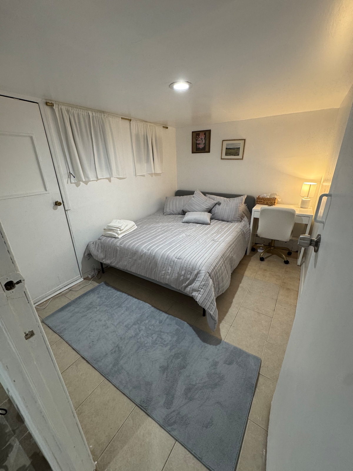 A cozy sleeping area is presented, featuring a spacious bed dressed in light gray striped bedding. A soft rug lies on the tile floor, while a small desk with a chair is positioned near a window, providing natural light. White curtains frame the window.