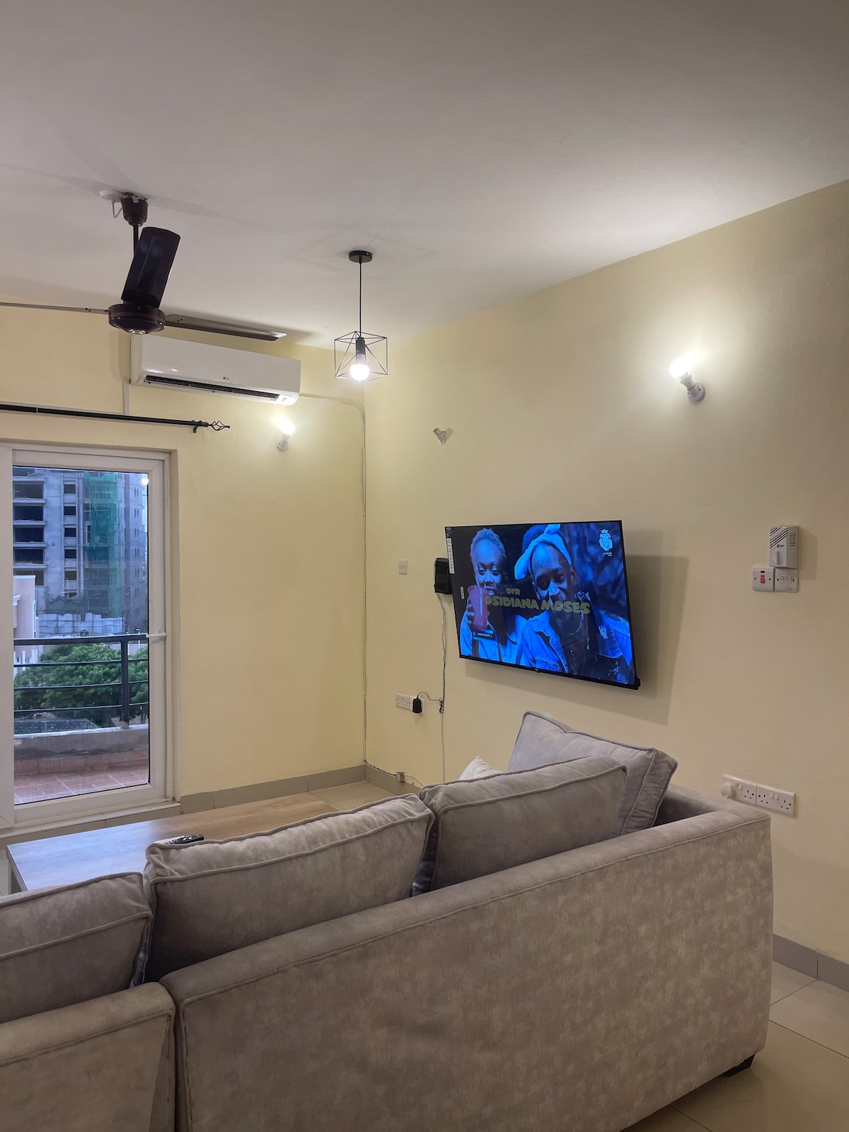 A comfortable living area features a light-colored sofa positioned towards a wall-mounted television. Soft lighting is provided by ceiling fixtures, while a sliding glass door leads to a balcony, allowing natural light to enter the space.
