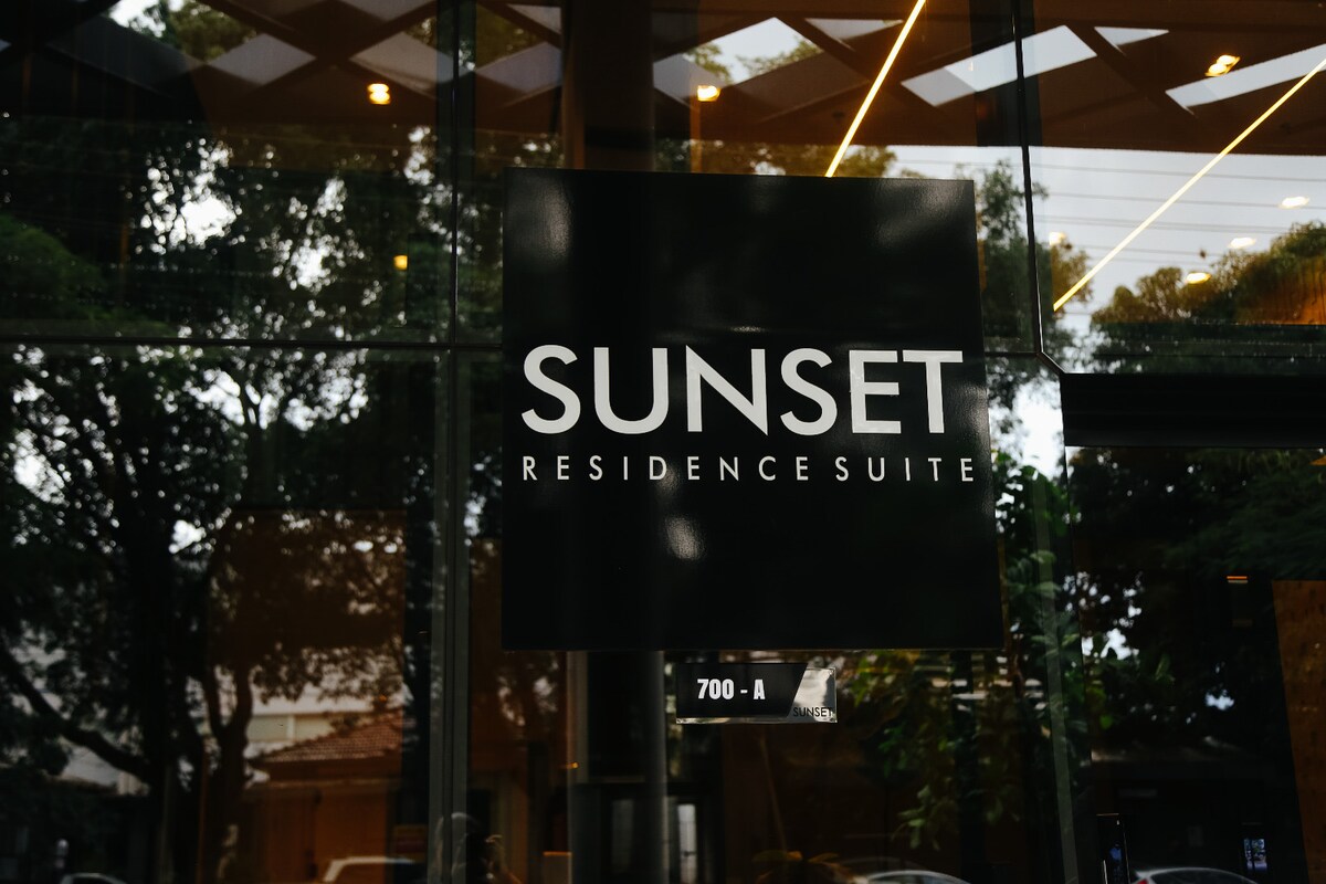 A modern sign displaying 'SUNSET RESIDENCE SUITE' is prominently featured. The surroundings are characterized by large glass windows that reveal greenery outside. Warm lighting reflects off the surfaces, creating a refined and welcoming entrance.