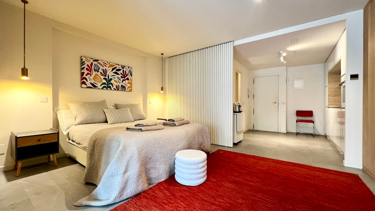 A spacious bedroom is lit by soft pendant lighting, featuring a comfortable bed with textured bedding. A colorful wall art piece adds character, while a plush round stool and a warm area rug complete the inviting atmosphere. A kitchenette is visible in the background.