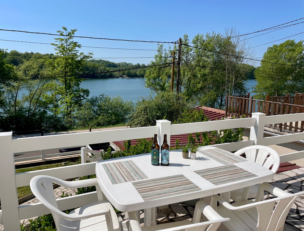 A serene outdoor space features a dining table with a striped runner, surrounded by white chairs. In the background, a calm lake is visible, framed by lush greenery and trees. Power lines and wooden structures can be seen in the distance, contributing to the natural setting.