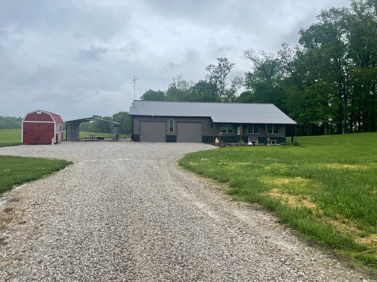 A gravel driveway leads to a single-story house with a metal roof, flanked by a spacious yard. A red barn is visible to the left, and a carport offers shaded parking. Surrounding greenery enhances the peaceful rural setting.